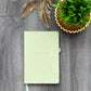 A pale sage green hardcover notebook with a subtle hexagonal pattern and the words 'Organise | Grow| Thrive' printed on the front, styled on a gray wood plank surface next to a potted green plant and dried palm leaves.